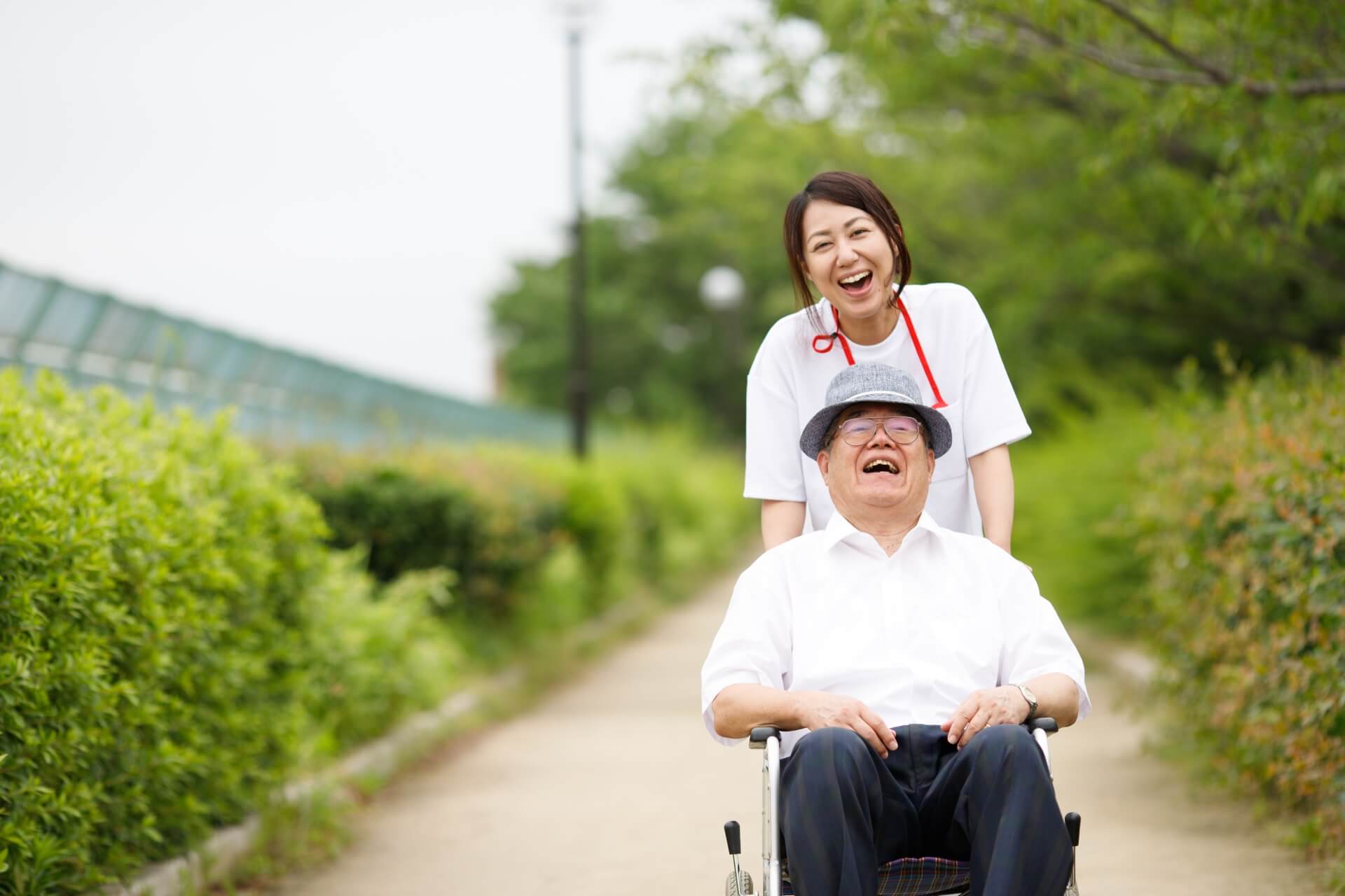 車椅子の高齢男性と、車椅子を押す女性介護スタッフが、外で笑っている写真