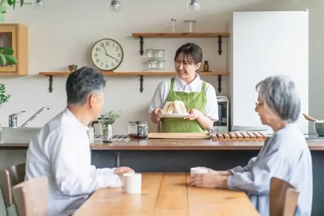 食卓に座る老夫婦に調理したサンドイッチを提供する、エプロン姿の女性家事代行スタッフの写真