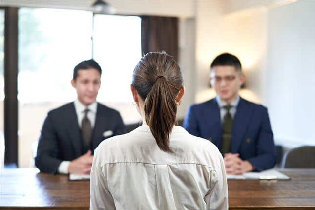 スーツを着た二人の男性と向き合い、机を挟んで会話している女性の後ろ姿の写真