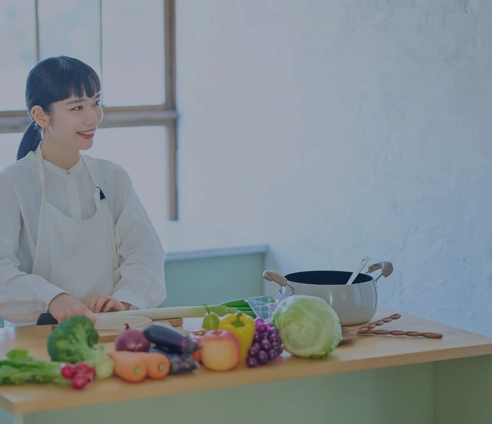 野菜や果物など、様々な食材が並べられた台所で、笑顔で調理している女性家政婦の写真