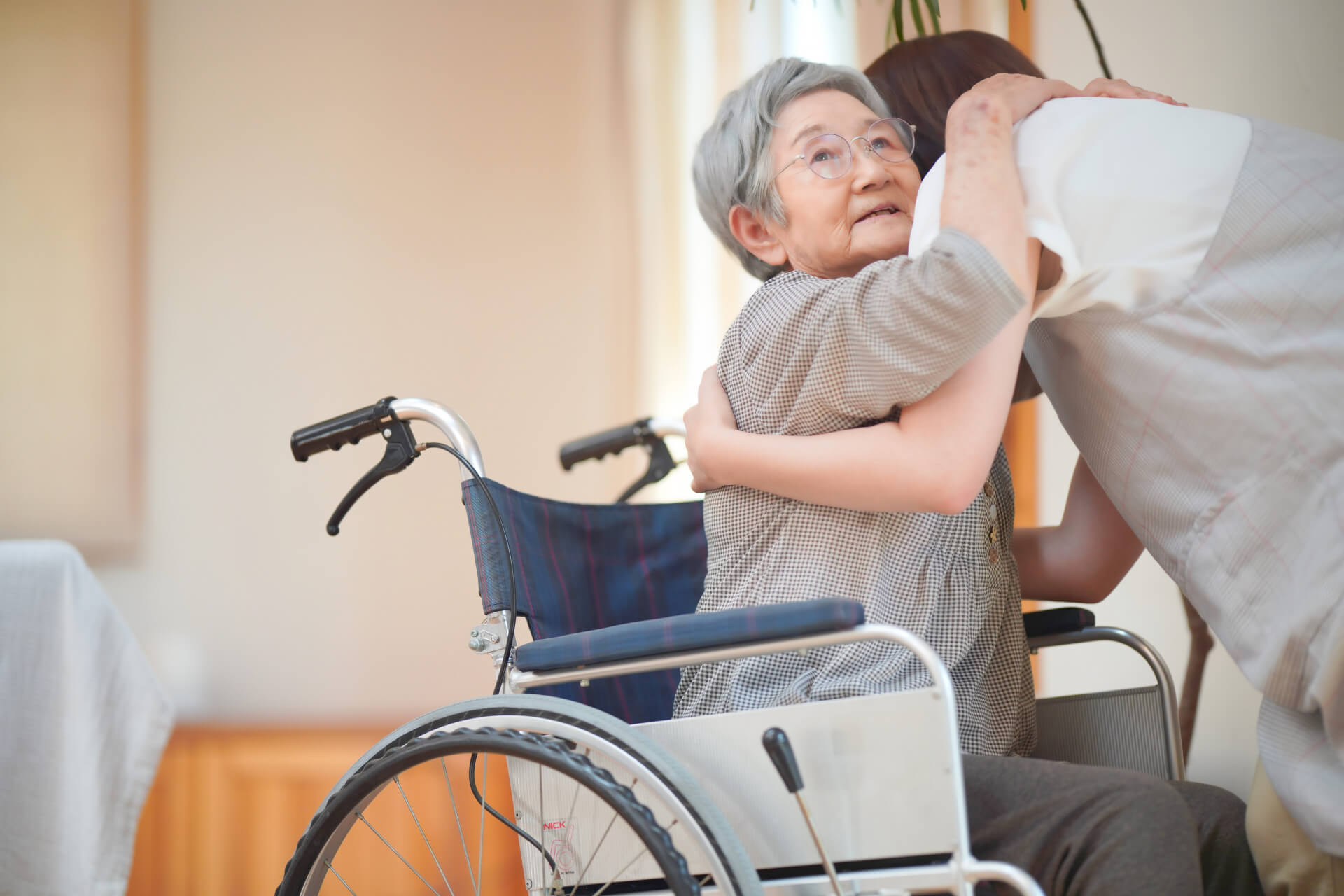 女性介護スタッフが、車椅子に乗った高齢女性が立ち上げるのを介助する写真