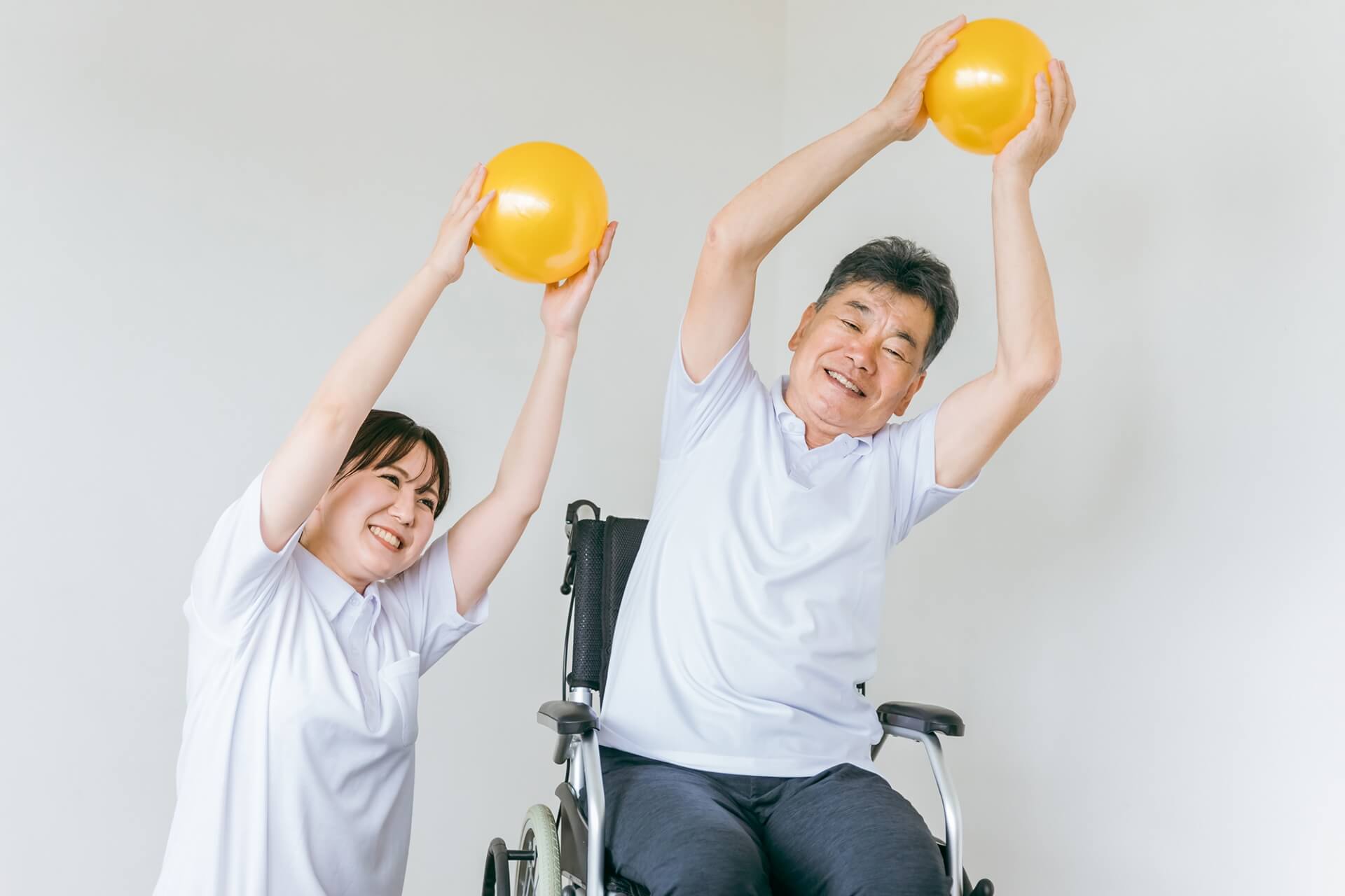 女性介護スタッフと、車椅子に乗ったご高齢の男性が、それぞれ両手で持ったボールを高く上げて運動している様子の写真