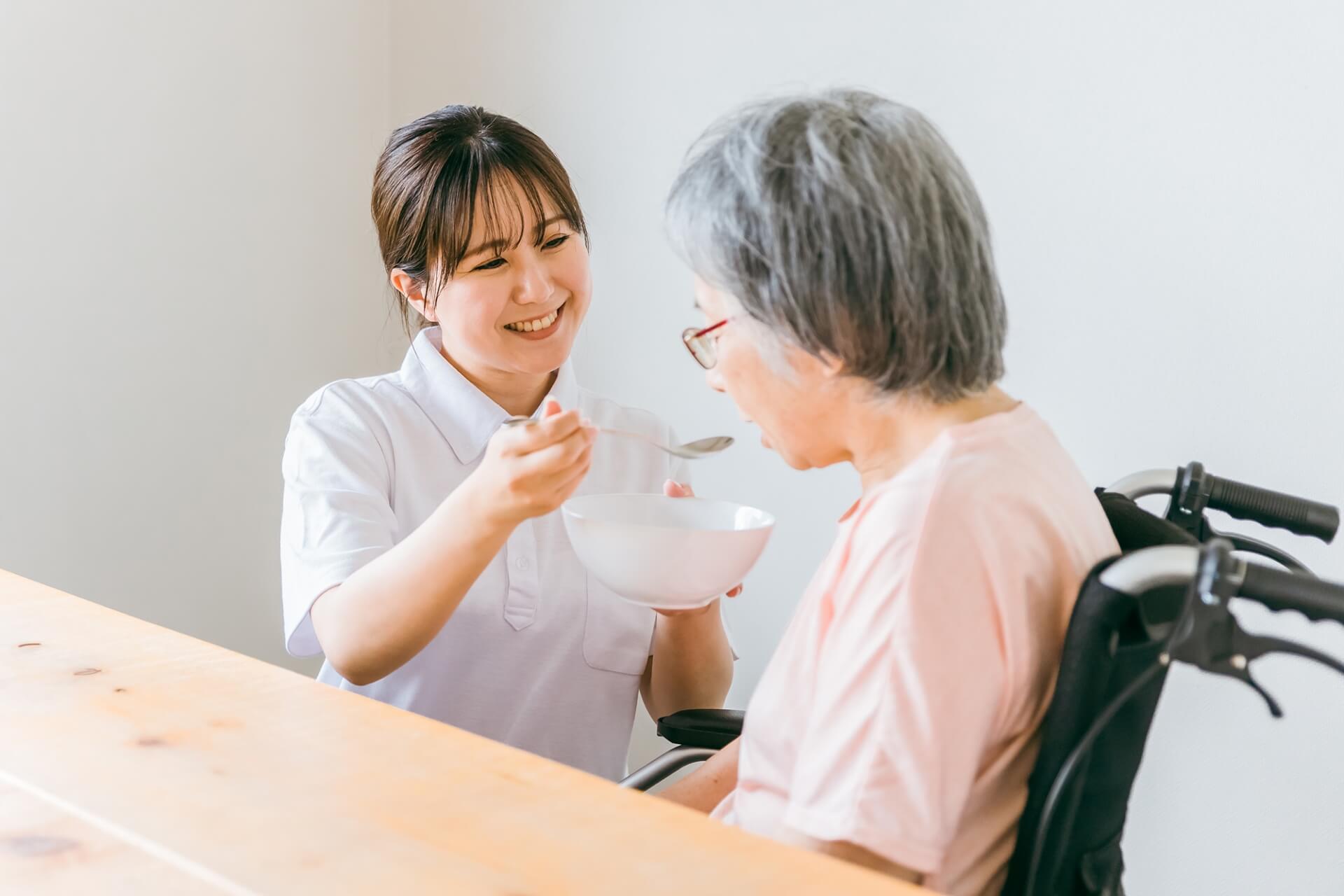 女性介護スタッフが、車椅子に乗った高齢女性の食事を笑顔で介助している写真