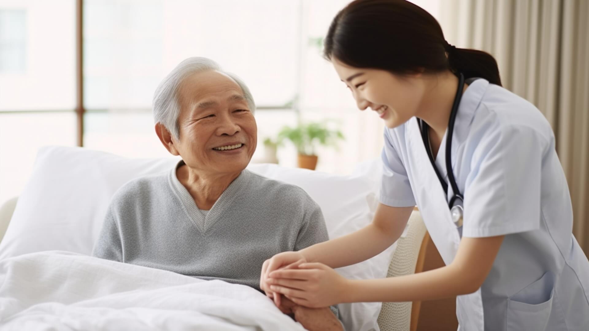 白衣を着た女性看護スタッフが、ベッドに入った白髪の高齢男性と親しく会話している写真