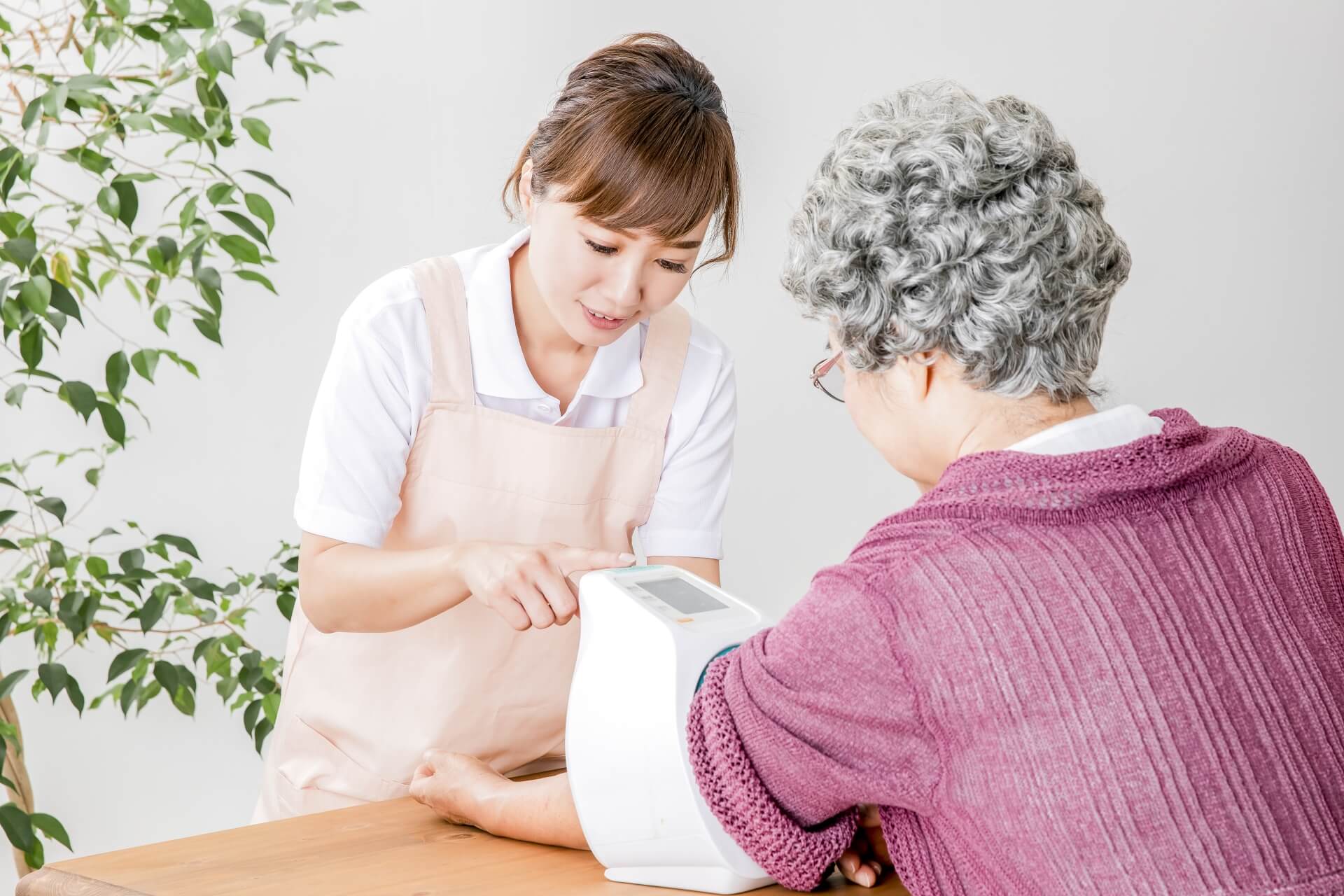 エプロンを身につけた明るい女性介護スタッフが、ご高齢女性の血圧を測定している写真