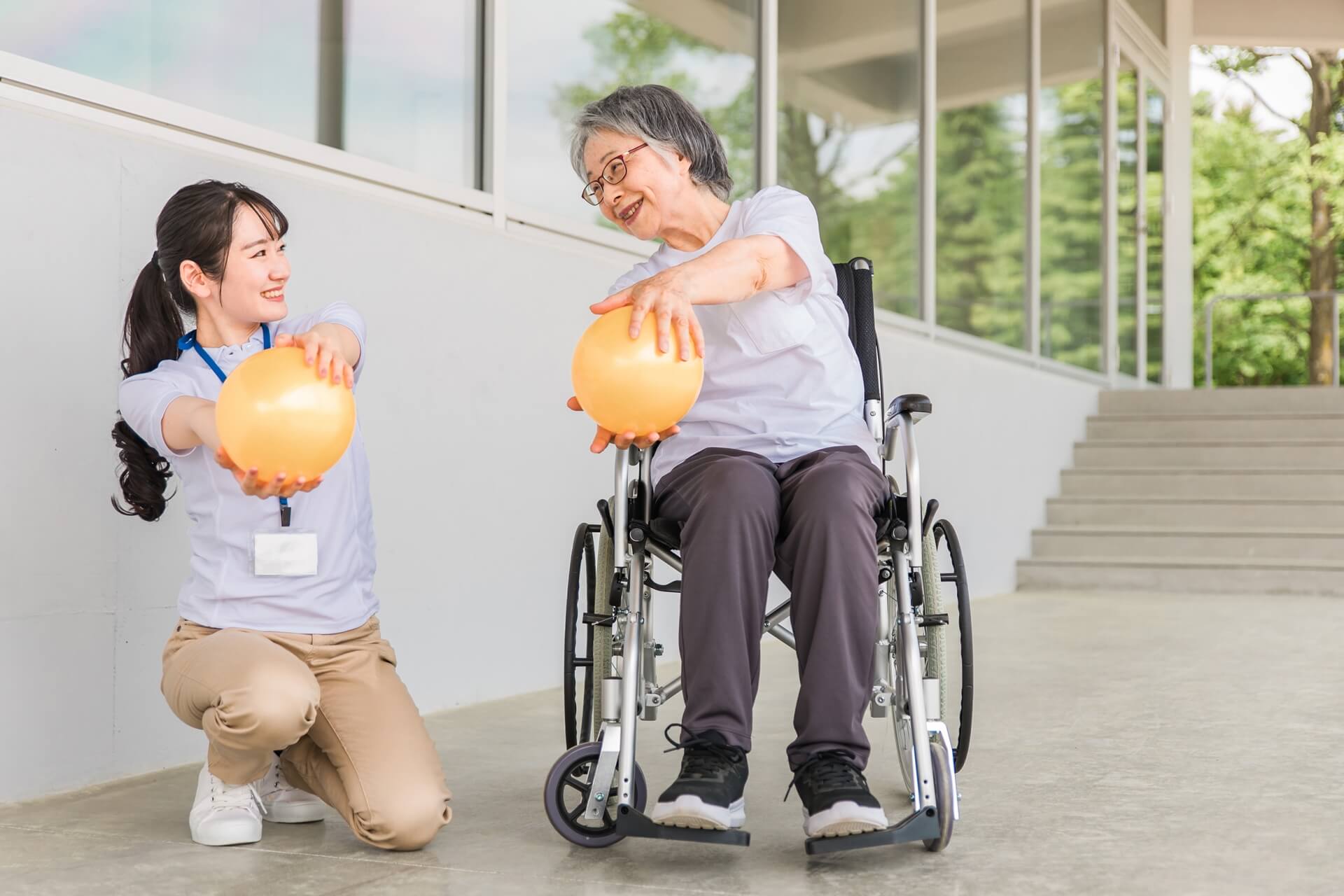 車椅子に乗った女性と女性介護スタッフが、空気の入ったボールを両手で持って、運動している様子の写真
