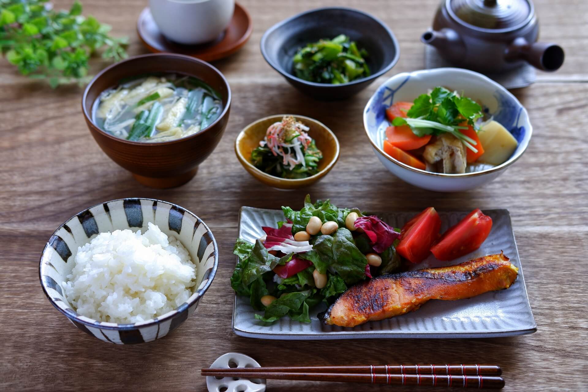 白米や味噌汁、魚、大豆サラダ、肉じゃが、漬物、ほうれん草、急須に入ったお茶、箸などが机に並べられた、日本の伝統的な朝食の写真